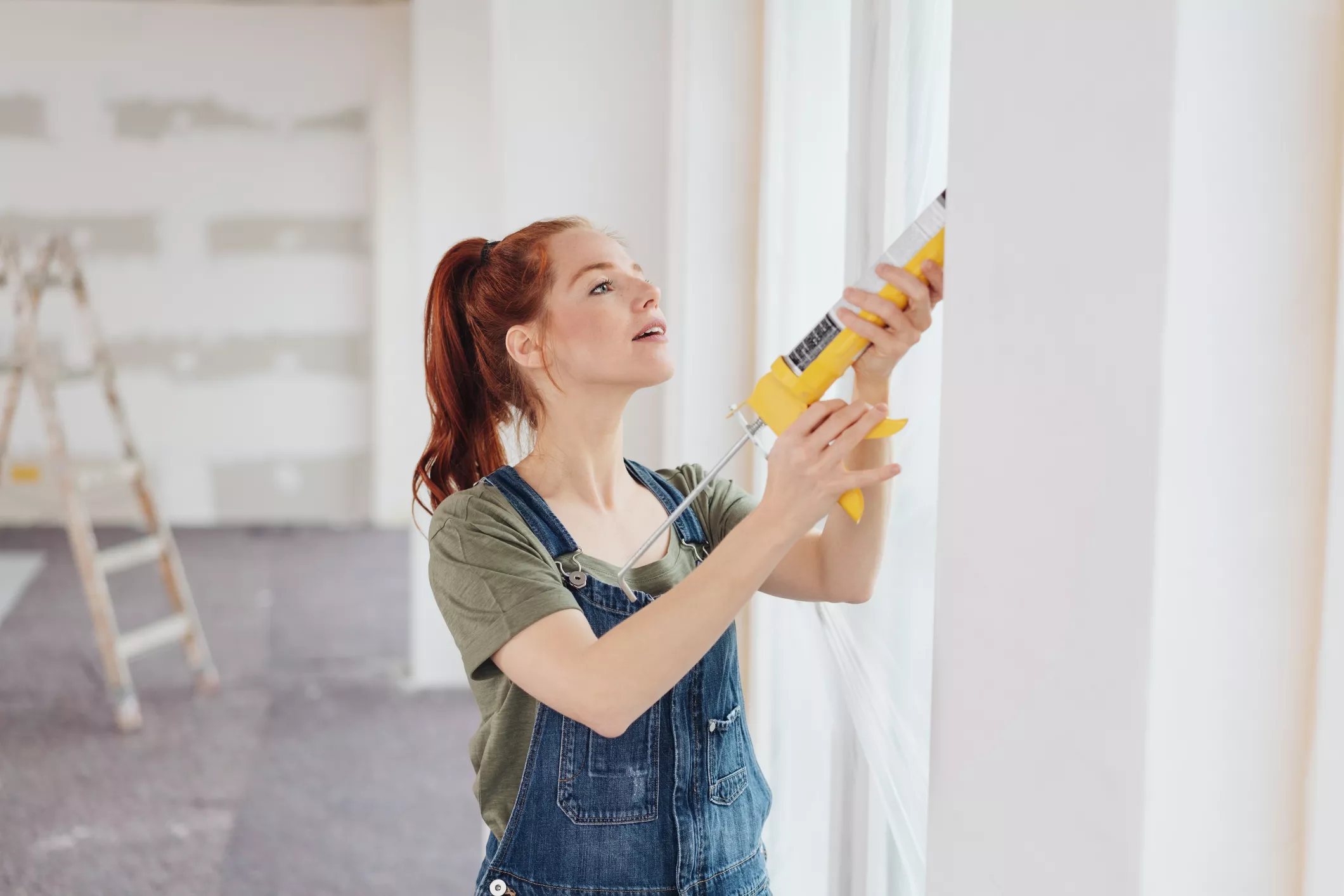 Young woman applying glue from a gun to a window frame during house renovation in an unfinished living room